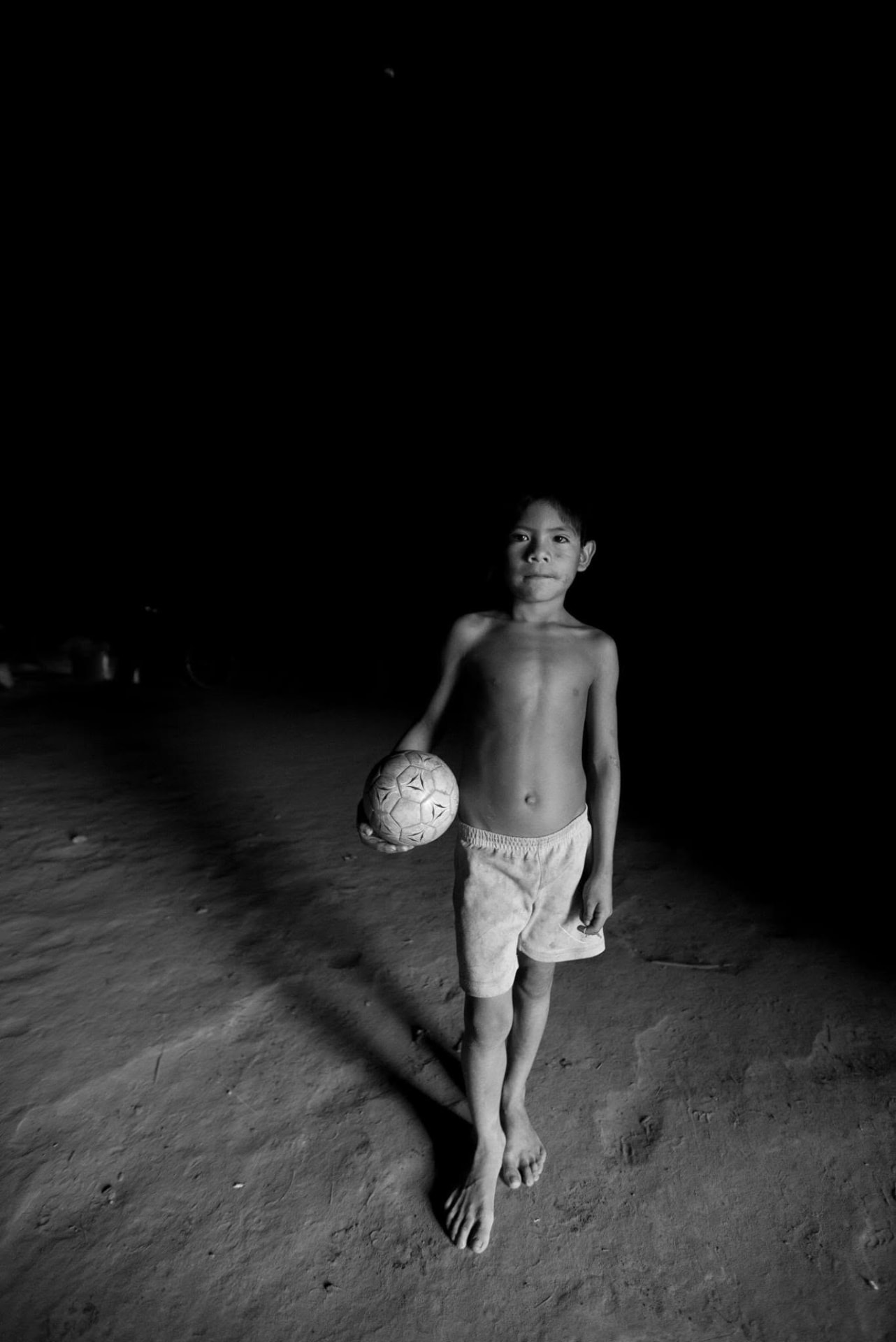 A barefoot boy holding a worn soccer ball against a black background