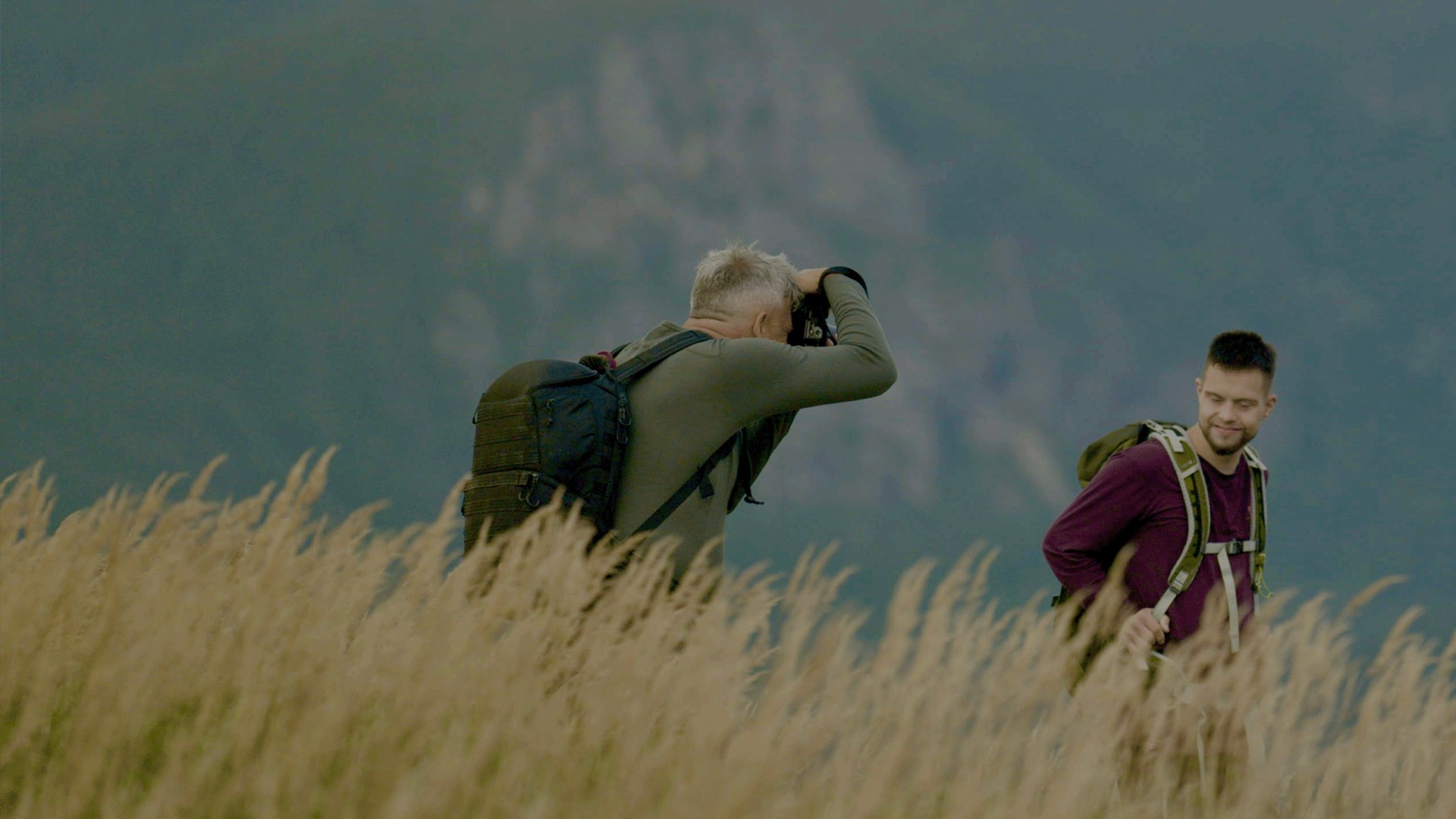 Two men hiking a grassy ridge, one filming