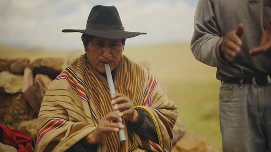 An Andean man in a black hat and striped poncho playing a flute