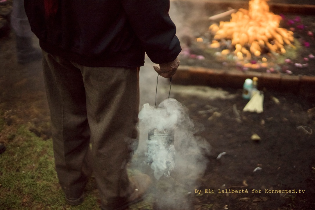 A person holding a smoking censer beside a ceremonial fire ringed with flowers and offerings
