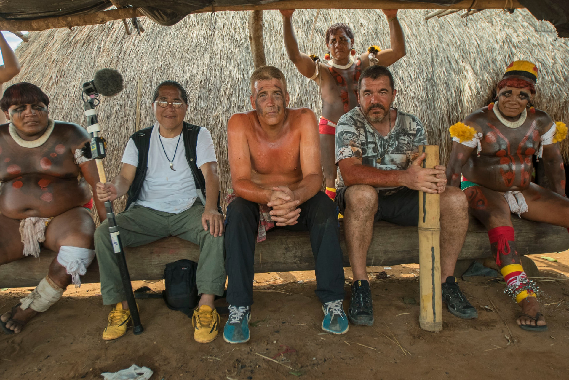 Éli and his crew seated with Kalapalo women in body paint under a thatched shelter